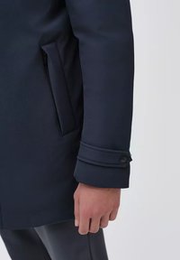 Cappotto blu navy in tessuto strutturato, con una tasca laterale e un dettaglio al polsino con bottoni. Il polsino presenta due cuciture visibili e una finitura liscia.