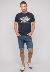 T-shirt en coton bleu marine avec un texte blanc de style vintage. Porté avec un short en denim et des chaussures à enfiler beige, avec des ourlets roulés.