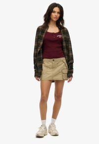 Brązowa koszula z flaneli w kratę na bordowym topie na ramiączkach, zestawiona z beżową spódnicą typu cargo z kieszeniami oraz beżowymi trampkami z białymi akcentami.