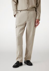 Beige cargobroek met een losse pasvorm, voorzien van zijzakken, een knoopsluiting en een gladde stoftextuur. Gdraag met zwarte instapschoenen.