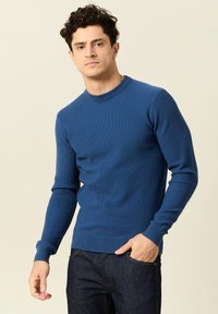 Blauwe geribbelde gebreide trui met lange mouwen en een ronde hals, gecombineerd met donkerdenim jeans. De stof heeft een gladde textuur en een getailleerd ontwerp.