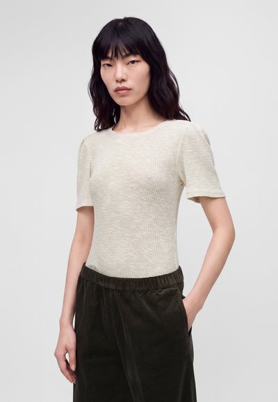 Femme aux longs cheveux noirs portant un haut crème texturé à manches courtes et un pantalon en velours côtelé vert foncé, debout avec une main dans la poche.