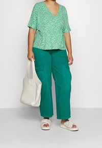 Top verde con estampado floral y escote en V, corte holgado; pantalones cargo anchos color teal con bolsillos laterales; bolso blanco; sandalias de crema con detalle anudado.