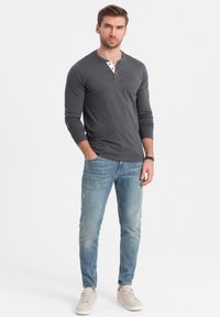 Grijze lange mouwen henley shirt met een knoopdetail, gecombineerd met lichtblauwe slim-fit jeans en lichte sneakers. Effen achtergrond.