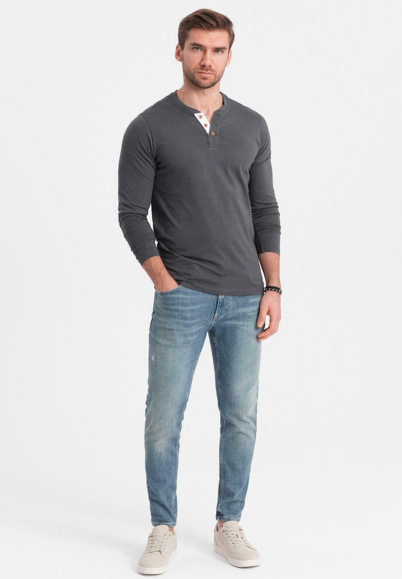 Grijze lange mouwen henley shirt met een knoopdetail, gecombineerd met lichtblauwe slim-fit jeans en lichte sneakers. Effen achtergrond.