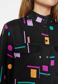 Černá košile s mandarínkovým límcem, zdobená barevnými geometrickými vzory v purpurové, modrozelené, oranžové a růžové barvě. Obsahuje dvě náprsní kapsy.