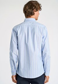 Camicia azzurro chiaro a righe verticali bianche, maniche lunghe e colletto classico. Presenta una piega sul retro e polsini con bottone.