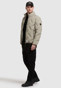 Uomo con giacca trapuntata beige, pantaloni cargo neri, stivali neri e cappellino da baseball nero, in piedi su uno sfondo semplice.