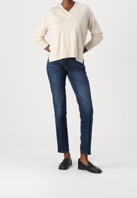 Beige v-neck villapaita pitkillä hihoilla, yhdistettynä tummansiniset suorat farkut ja mustat loafer-kengät, seisoo yksinkertaisella taustalla.