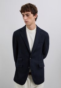 Joven con blazer azul marino oscuro sobre un cuello de tortuga blanco, de pie con las manos detrás de la espalda, mirando hacia abajo contra un fondo gris liso.