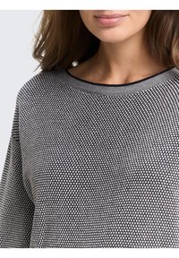 Femme portant un haut tricoté à motif noir et blanc avec un col rond, montrant ses lèvres et une partie de ses cheveux.