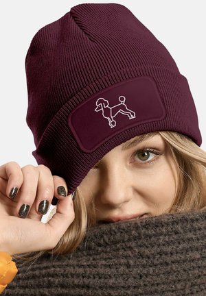 Burgundy knit beanie with a soft texture, featuring a patch displaying a white outline of a poodle. Worn slightly askew over a scarf.
