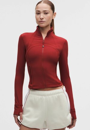 Žena má na sebe priliehavú červenú bundu na zips s dlhými rukávmi a biele vysokopásové šortky s bočnými vreckami, stojí pred jednofarebným pozadím.