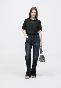 Μαύρο βαμβακερό t-shirt με λογότυπο, συνδυασμένο με σκούρο μπλε τζιν παντελόνι καμπάνα. Το μοντέλο κρατά μια υφασμάτινη πράσινη τσάντα και φοράει μαύρα σανδάλια.