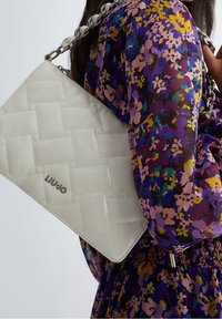 Nainen, jolla on päällään violetti kukallinen mekko, kantaa valkoista tikattua olkalaukkua, jossa on paksu ketjuhihna ja Liu Jo -logo.