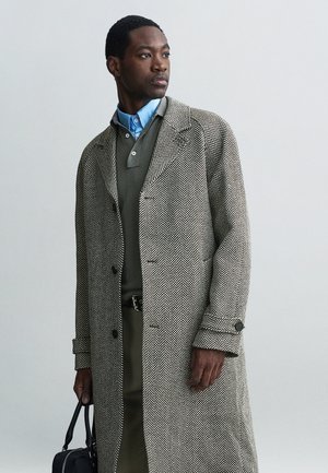Hondstooth-patroon overjas in grijs en wit, gecombineerd met een groene trui en een blauwe overhemd. Accessoires omvatten een zwarte tas.