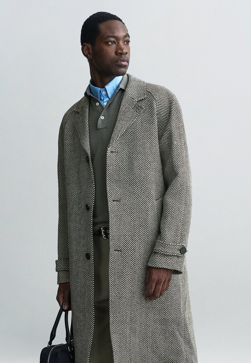 Kaput s uzorkom houndstooth u sivoj i bijeloj boji, uparen s zelenim duksericom i plavom košuljom s kragnom. Dodaci uključuju crnu torbu.