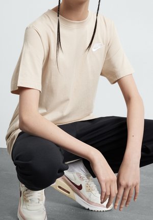 Person, der sidder på hug iført en beige Nike t-shirt, sorte bukser, hvide sokker og hvide Nike sneakers med rød swoosh.