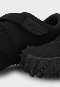 Zapatillas deportivas negras hechas de malla y materiales sintéticos, con una superficie texturizada, picos de goma en la suela y una correa con marca.