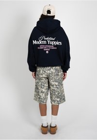 Personne portant un sweat à capuche noir avec le texte « Modern Yuppies », un short camouflage, des chaussettes blanches, des chaussures marron et une casquette beige, vue de dos sur un fond gris.