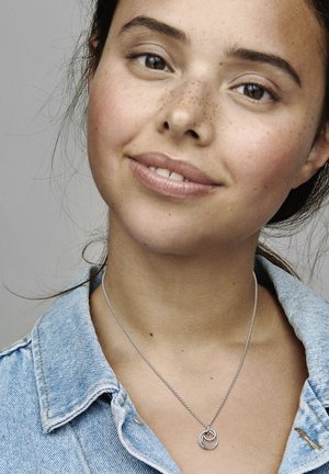 Collana in argento con pendenti circolari intrecciati. Indossata sopra una camicia di denim, evidenziando una consistenza e una posizione casual.