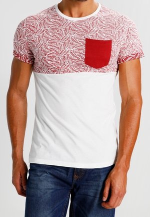Homme portant un T-shirt blanc et rouge à manches courtes avec un motif de feuilles sur la moitié supérieure et la moitié inférieure unie, associé à un jean bleu.