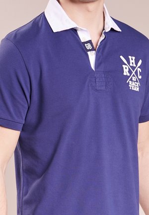 Granatowa koszulka polo z białym kołnierzykiem, z haftowanym logo i wzorem skrzyżowanych wioseł. Wykonana z gładkiego materiału, z krótkim rękawem.