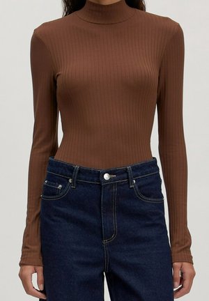 Person iført en tætsiddende brun ribbet langærmet rullekravtrøje, der er stoppet ned i højttaljede mørkeblå jeans med knaplukning.