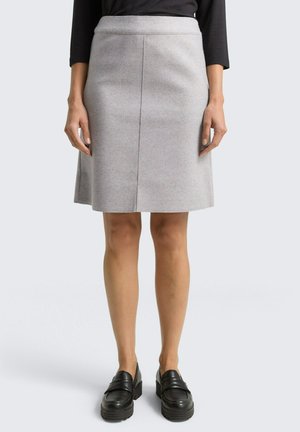 Frau trägt einen knielangen hellgrauen Rock, ein schwarzes Top und schwarze klobige Loafers, steht vor einem einfarbigen Hintergrund.