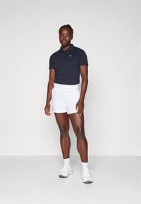 Camisa polo azul-marinho com colarinho e mangas curtas, combinada com calções de atletismo brancos. O modelo usa ténis brancos e está numa fundo neutro.