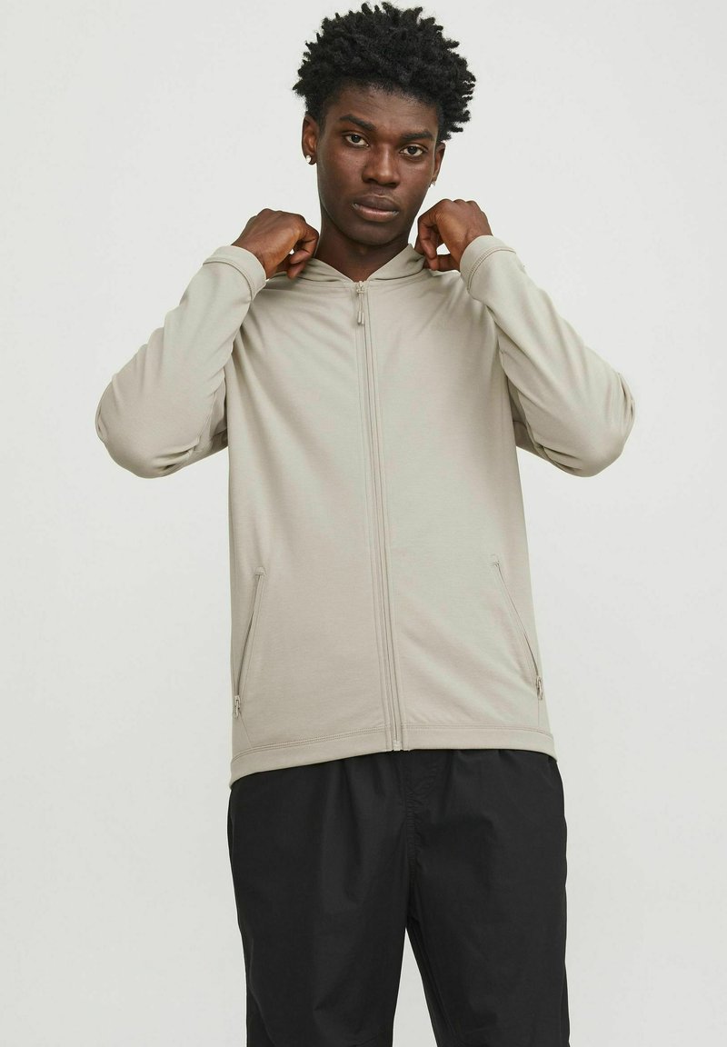 Light beige zip-up hoodie with a hood, two side pockets, and long sleeves, worn over black pants. Smooth texture, casual design.
