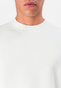 Primer plano de la parte inferior de la cara y el cuello de una persona que lleva un suéter básico de cuello alto blanco contra un fondo blanco.