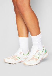 Baskets blanches avec des accents roses et verts, en mesh et matériau synthétique, avec un design à lacets, présentant une semelle texturée et un détail de logo.