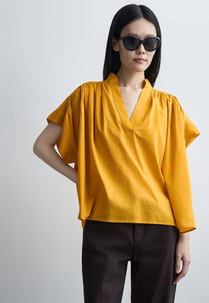 Žena s čiernymi slnečnými okuliarmi, voľným horčicovožltým topom s krátkymi a dlhými asymetrickými rukávmi a čiernymi nohavicami na jednotnom pozadí.