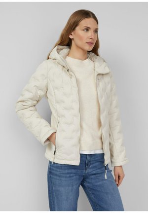 Kobieta ubrana w kremową pikowaną kurtkę z kapturem, nałożoną na biały sweter i niebieskie dżinsy, stojąca z jedną ręką w kieszeni, patrząca w prawo.