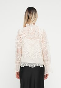 Blusa blanca transparente con patrón de encaje floral, cuello acampanado y mangas acampanadas, combinada con una falda negra lisa. Detalles de diseño texturizado visibles.