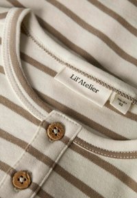 Koszula w paski w kolorze brązowym i białym z okrągłym dekoltem, drewnianymi guzikami i metką z napisem "Lil' Atelier". Wykonana z miękkiej bawełny.