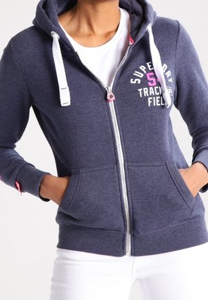 Sweat à capuche bleu marine zippé avec cordons blancs et touches roses, porté sur une chemise blanche, mains dans les poches avant.