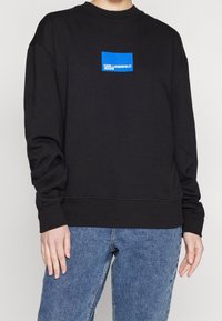 Sudadera negra de manga larga, con cuello redondo y un logo rectangular azul en el frontal. Fabricada con un tejido suave y de corte relajado.