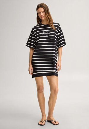 Vrouw die staat in een zwart-wit gestreepte oversized T-shirtjurk met de tekst ""Jack Jeans"", draagt zwarte slippers, neutrale achtergrond.