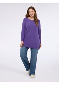 Maglione viola a maglia con trama a coste e spacchi laterali, abbinato a jeans a gamba larga azzurri e sneaker crema, su sfondo neutro.