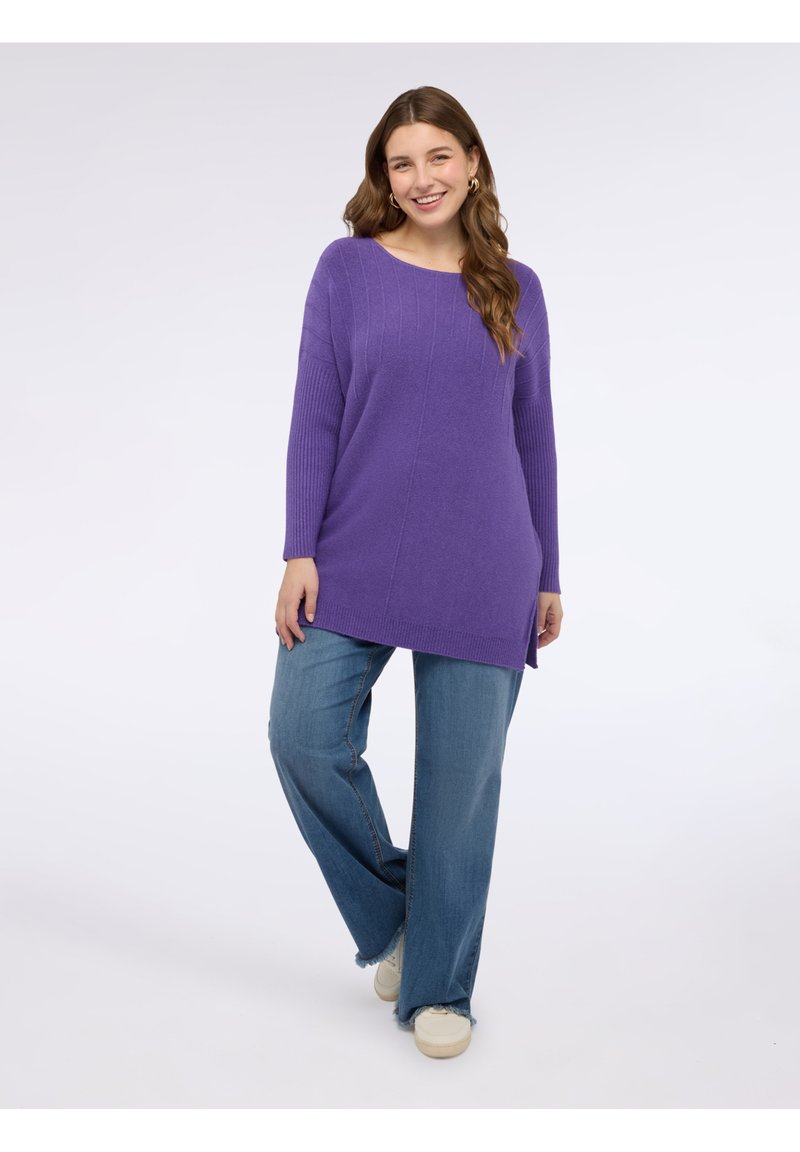 Maglione viola a maglia con trama a coste e spacchi laterali, abbinato a jeans a gamba larga azzurri e sneaker crema, su sfondo neutro.