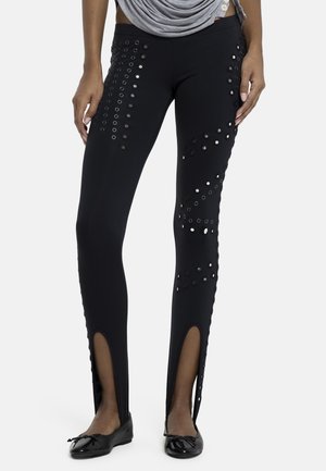 Svarte tettsittende bukser med hullmønster på lår og legger, frontsplitt ved anklene, brukt med svarte ballettsko og en grå rynket topp.