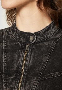 Svart denimjacka med hög krage, silverfärgad knäppning och guldfärgad dragkedja. Tyget har en subtil textur med en sliten look.