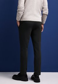 Svarta slim-fit byxor med sidofickor, kombinerade med en ljusgrå strukturerad tröja och svarta snörskor, mot en blå bakgrund.