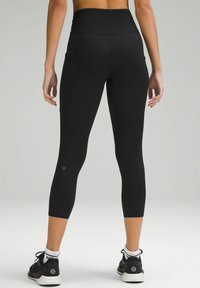 Leggings capri negre cu talie înaltă, textură netedă și două buzunare laterale; asortate cu pantofi sport negri ce au accente albe.