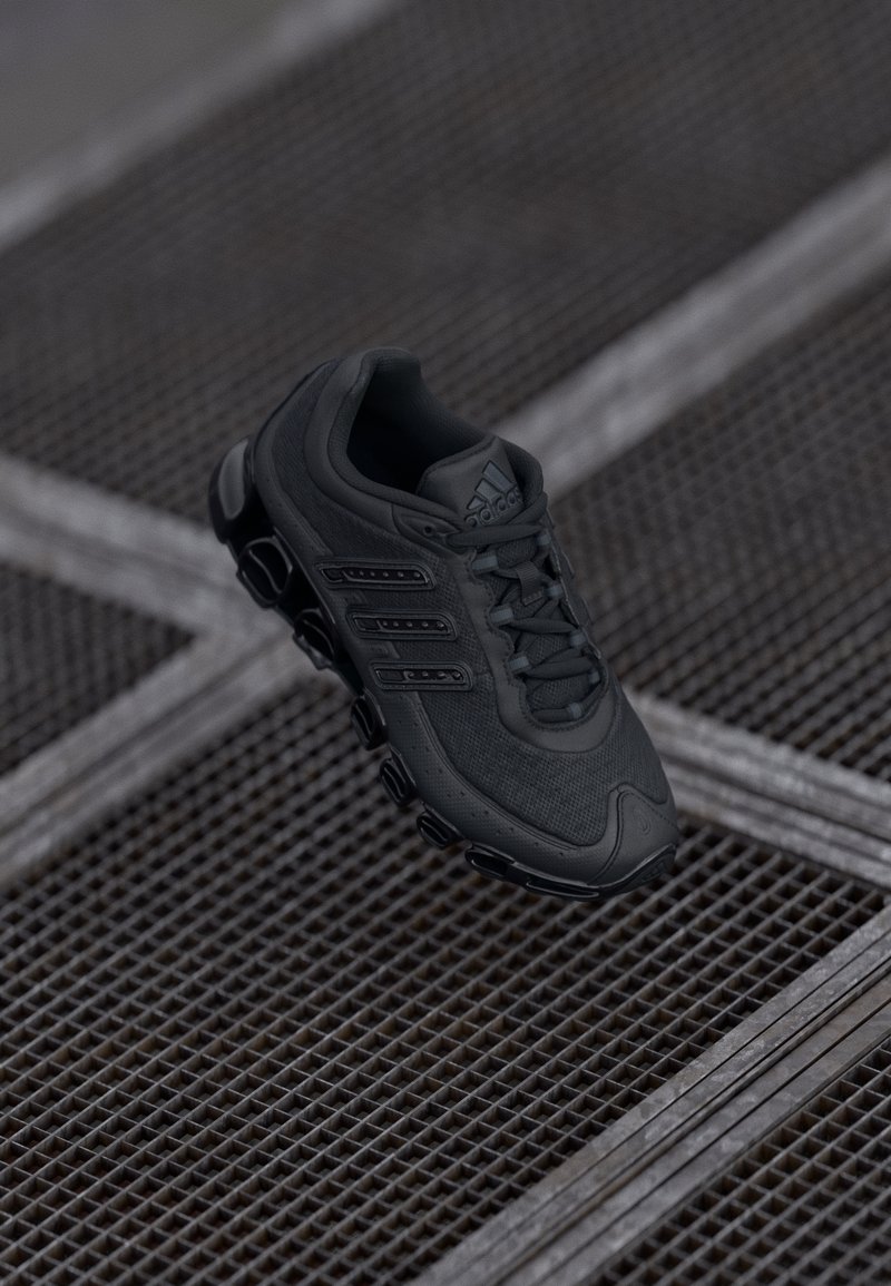 Černá běžecká bota Adidas s texturovanou podrážkou a třemi pruhy, umístěná nad kovovou mřížkou s protínajícími se čarami.