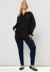 Schwarze Bluse mit Knöpfen und strukturiertem Karomuster sowie Rüschensaum, kombiniert mit dunkelblauen Skinny-Jeans und schwarzen Ballerinas.