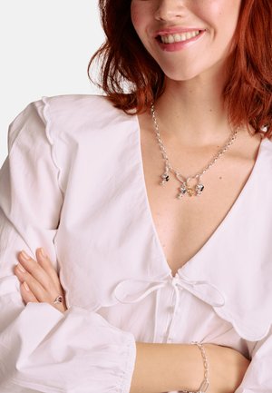 Silberne Halskette mit Anhängern, runde Glieder, gemischte Farben, getragen über einer weißen Bluse mit geschwungenem Ausschnitt und einer gebundenen Schleife. Glatte Textur.