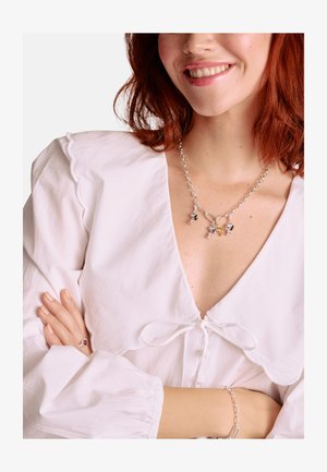 Silberne Halskette mit Anhängern, runde Glieder, gemischte Farben, getragen über einer weißen Bluse mit geschwungenem Ausschnitt und einer gebundenen Schleife. Glatte Textur.